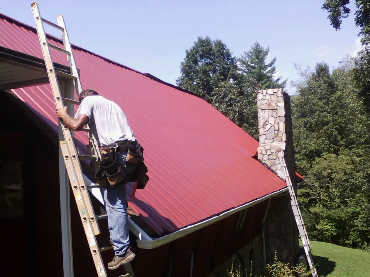 Roofer on a ladder beside a red metal roof and stone chimney — Soffit & Fascia Repair by Roofing Brothers in Middlebury